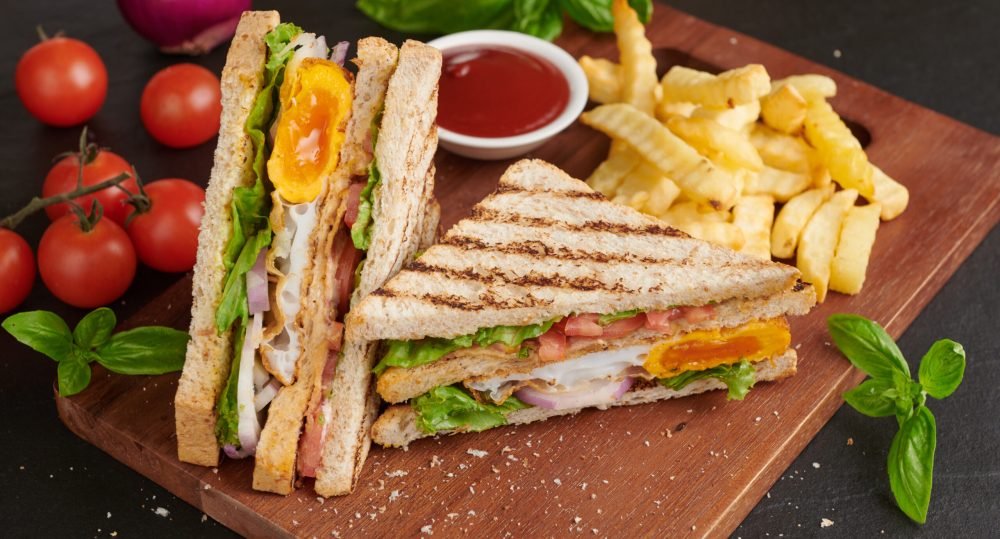 Club-sandwich with french fries. Sandwich with bacon, fried egg. Grilled and pressed toast with bacon, fried egg, tomato and lettuce served on wooden cutting board.