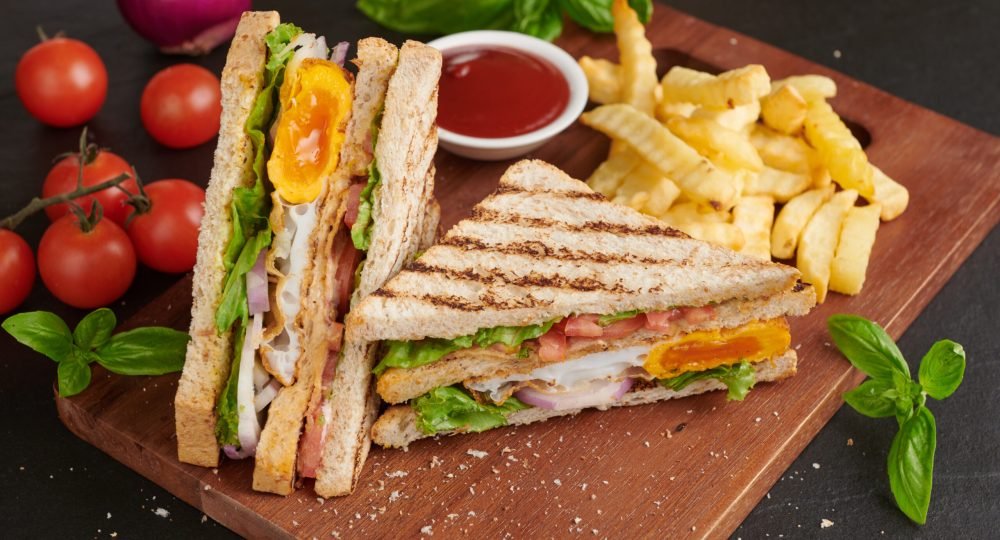 Club-sandwich with french fries. Sandwich with bacon, fried egg. Grilled and pressed toast with bacon, fried egg, tomato and lettuce served on wooden cutting board.